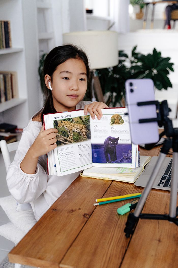 services-05 Young girl displays book during online class using smartphone and tripod.