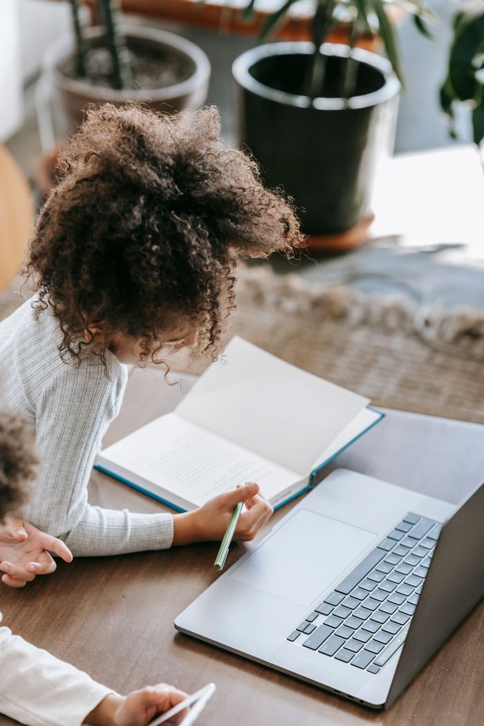 From above of unrecognizable African American schoolgirl with curly hair in casual clothes lying on floor and taking notes in copybook during distance lesson via laptop at home